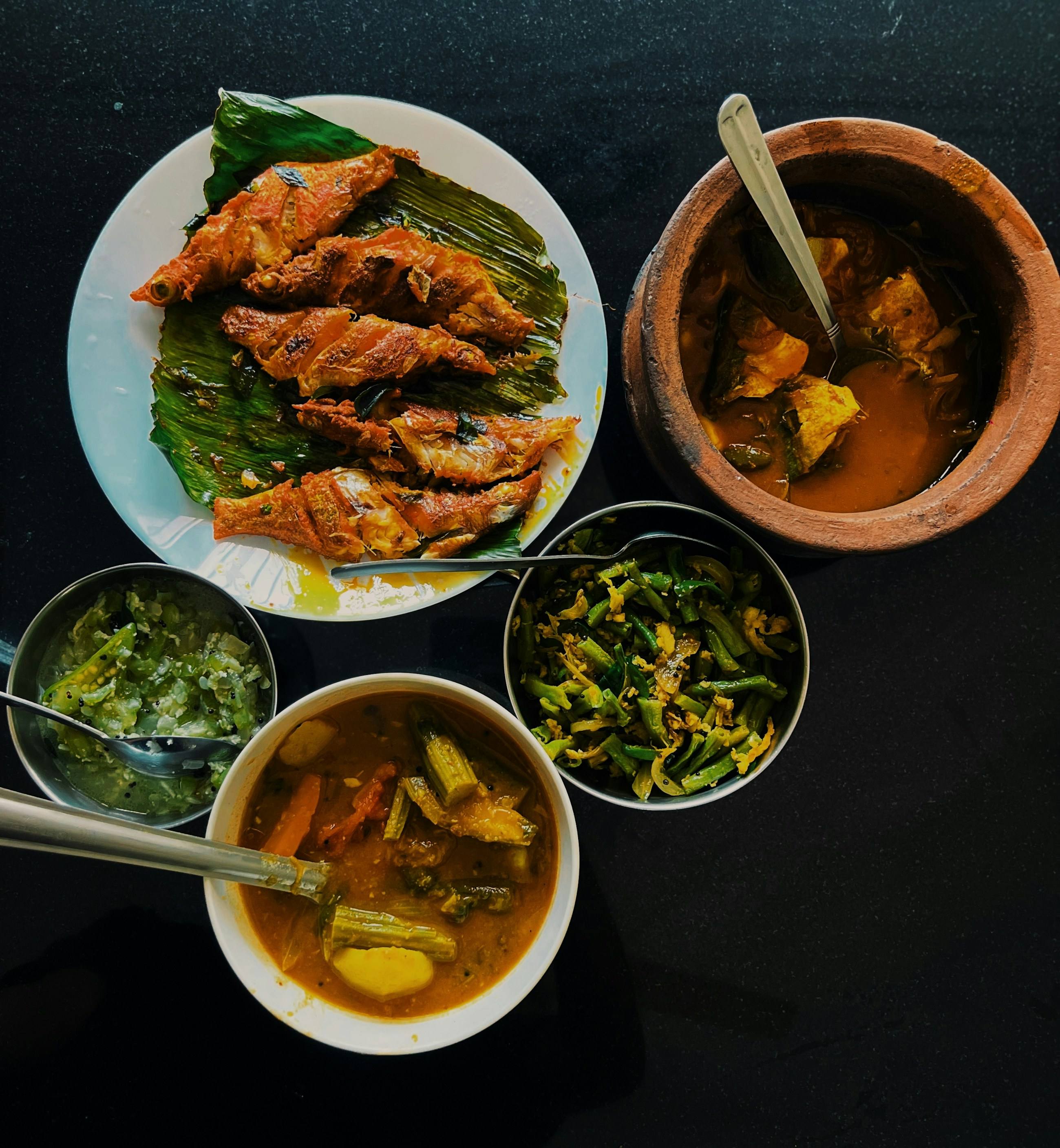 A Kerala meal from above: fried fish on a banana leaf, fish curry in a clay pot, bean thoran and sambar on a black table
